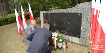 Park. Przed pomnikiem ofiar katyńskich dwóch mężczyzn w średnim wieku kuca. Przy pomniku leży wiązanka z białymi kwiatami i stoją trzy znicze. Po obu stronach pomnika stoją na stojaku trzy biało-czerwone flagi.