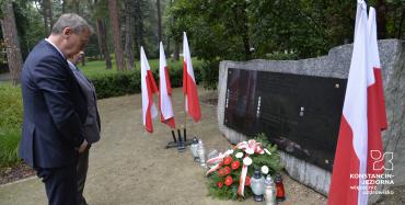 Park. Przed pomnikiem ofiar katyńskich stoi dwóch mężczyzn w średnim wieku. Przy pomniku stoją znicze i leży wiązanka z białymi kwiatami. Po obu stronach pomnika stoją trzy biało-czerwone flagi.