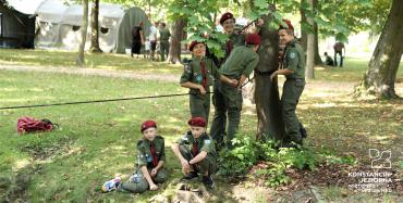 Park Zdrojowy. Grupa sześciu chłopców stoi obok siebie przy drzewie. Do drzewa przywiązana jest lina. Wszyscy są ubrani w harcerskie stroje, na głowach mają bordowe berety. 