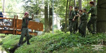 Park Zdrojowy. Chłopiec stoi na linie przywiązanej do drzewa. Obok niego na trawie stoi pięć osób. Wszyscy są ubrani w harcerskie stroje.