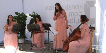 Pod namiotem na krzesłach siedzą trzy młode kobiety. W rękach trzymają skrzypce, przed nimi stoją na stojaku kartki z nutami. Każda ma na sobie różową sukienkę. Jedna z kobiet w ręku trzyma mikrofon.