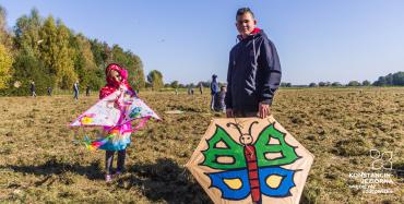 Łąki Oborskie. Chłopiec i dziewczynka w wieku szkolnym stoją obok siebie. Dziewczynka trzyma w rękach różowy latawiec w kształcie trójkąta. Chłopiec trzyma w ręku latawiec w kształcie sześciokąta , na którym namalowany jest kolorowy motyl. W tle drzewa i błękitne niebo.