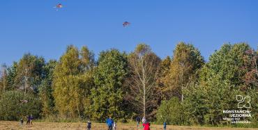 Łąki Oborskie. Dziesięć osób – dorośli i dzieci puszczają na błękitnym niebie latawce. W tle zielone drzewa. 