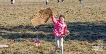 Łąki Oborskie. Dziewczynka w wieku szkolnym stoi na trawie, w ręku trzyma latawiec w kształcie sześciokąta. Do latawca przywiązany jest sznurek. W tle jest sześć osób z latawcami.