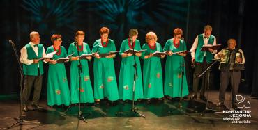 Na scenie występuje dziewięć osób. Przed nimi stoją mikrofony. Obok siebie stoi sześć kobiet i trzech mężczyzn. Kobiety w rękach trzymają śpiewniki, ubrane są w zielone sukienki, na szyi mają czerwone korale. Jeden z mężczyzn gra na akordeonie.