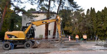 Remont drogi. Widać rozkopaną ziemię, obok stoi żółta koparka. Przy koparce stoi dwóch robotników, którzy ubrani są w pomarańczowe kurtki z odblaskami.