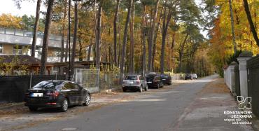 Ulica z kostki brukowej. Po jej lewej stronie stoją zaparkowane w rzędzie samochody. W głębi widać duży budynek. Po prawej stronie ulicy znajduje się chodnik. W dali widać wysokie drzewa z żółtymi i brązowymi liśćmi. 