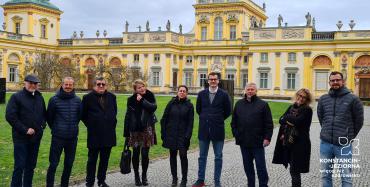 Dziewięć osób stoi obok siebie w rzędzie na chodniku. W tle jest budynek Muzeum Pałacu Króla Jana trzeciego w Wilanowie.