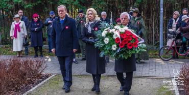 Park. Dwóch mężczyzn i jedna kobieta stoją obok siebie w rzędzie. Jeden z mężczyzn w rękach trzyma duży wieniec z biało-czerwonymi kwiatami.