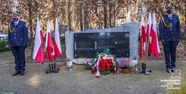 Przy pomniku Pamięci Ofiar Katynia stoi dwóch mężczyzn, którzy ubrani są w mundury Straży Miejskiej. Obok pomnika ustawione są biało-czerwone flagi oraz stoją kwiaty i znicze.