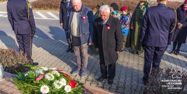 Trzech mężczyzn w średnim wieku stoi przy pomniku. Na pomniku leży wiązanka z biało-czerwonymi kwiatami.