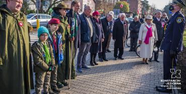 Około piętnastu osób stoi obok siebie w rzędzie na chodniku. W tle widać ulicę i budynki. 