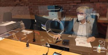 Przy biurku na krześle siedzi kobieta, wzrok ma skierowany na ekran komputera. Na twarzy ma maseczkę ochronną, na nosie okulary. Przed nią stoi drugie biurko. Biurka oddzielone są przezroczystą osłoną z tworzywa sztucznego. 