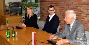 Sala konferencyjna. Trzy osoby siedzą na krzesłach przy drewnianym stole. Mężczyzna w średnim wieku, młody mężczyzna oraz młoda kobieta. Przed nimi stoją dwa mikrofony, butelki z wodą oraz mała flaga Polski i flaga Włoch. W tle ściana z czerwonej cegły.