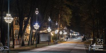 Aleja spacerowa nocą. Po lewej stronie widoczne są latarnie, na których zamontowano zwisające ozdoby świąteczne.
