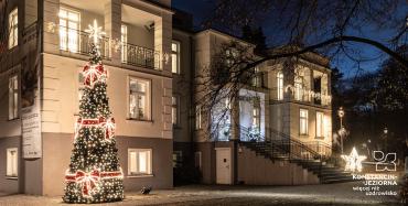  Budynek, który jest oświetlony nocą. Po lewej stronie stoi duża choinka ozdobiona czerwoną kokardką i lampkami. 