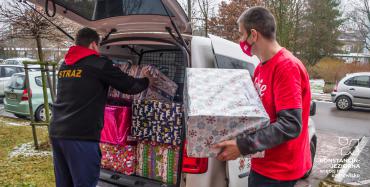 Samochód dostawczy, który ma otwarty bagażnik. Znajdują się w nim duże pudła – prezenty świąteczne. Obok samochodu stoi dwóch mężczyzn, jeden pokuje do bagażnika pudełka, drugi mu je podaje. 