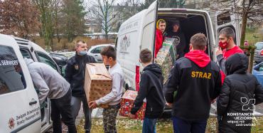 Dwa samochody dostawcze stoją obok siebie. Mają otwarte bagażniki. 9 osób, przepakowuje duże pudła – prezenty świąteczne z jednego samochodu do drugiego. 