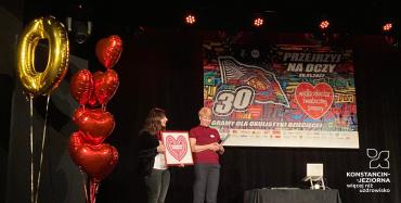 Na scenie występują 2 kobiety. Kobieta pierwsza prezentuje obraz z czerwonym sercem, druga trzyma w ręce mikrofon. Scena ozdobiona czerwonymi balonami w kształcie serc po lewej stronie oraz kolorowym banerem z tyłu. 