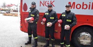 Czerwony autobus. Obok niego stoi 3 mężczyzn – strażaków w mundurach. W rękach trzymają czerwone koce. 