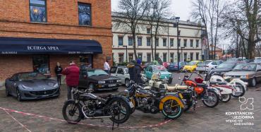 Parking z kostki brukowej. Na pierwszym planie  7 zabytkowych motocykli w głębi 4 zabytkowe samochody. Pomiędzy nimi oglądający ludzie. W tle budynki oraz parking ze współczesnymi samochodami.