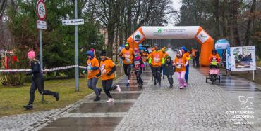 Brukowana aleja w parku. Na pierwszym planie biegnie grupa osób ubranych w pomarańczowe koszulki. Za plecami mają dmuchaną bramę w kolorze pomarańczowym – stanowiącą linię startu biegu. W tle liczne drzewa.