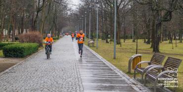 Brukowana aleja w parku. Po bokach ławki. Aleją biegnie grupa osób w pomarańczowych koszulkach a  jedna osoba jedzie na rowerze. W tle liczne drzewa.