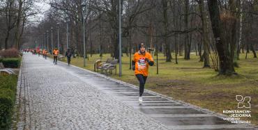 Brukowana aleja w parku. Po bokach ławki. Aleją biegnie kobieta w pomarańczowej koszulce a w tyle w rzędzie biegnie grupa podobnie ubranych ludzi. W tle liczne drzewa.