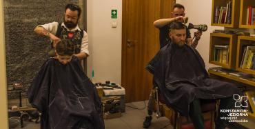 Salon fryzjerski. Na pierwszym planie w fotelach siedzą: chłopiec w wieku szkolnym i mężczyzna z brodą. Za nimi stoją fryzjerzy wykonując czynności pielęgnacyjne włosów głowy.
