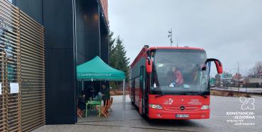 Na chodniku stoi duży czerwony autobus z białym napisem z boku Mobilny Punkt Poboru Krwi. Obok niego stoi zielony namiot. 