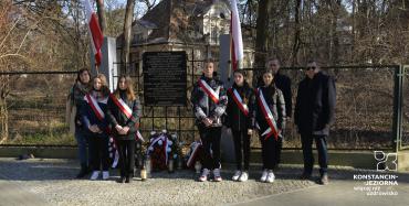 Pośrodku widoczna tablica pamiątkowa umieszczona w ogrodzeniu, za ogrodzeniem widoczny zabytkowy niski budynek. U góry ogrodzenie, po bokach tablicy dwie flagi biało-czerwone, przed tablicą leżą kwiaty i stoją znicze. Wzdłuż ogrodzenia stoi piątka nastolatków przepasanych z szarfami biało-czerwonymi, obok nich po prawej dwóch mężczyzn i po lewej kobieta