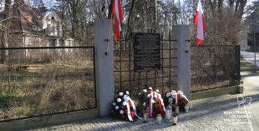 Pośrodku widoczna tablica pamiątkowa umieszczona w ogrodzeniu, za ogrodzeniem widoczny zabytkowy niski budynek. U góry ogrodzenie, po bokach tablicy dwie flagi biało-czerwone, przed tablicą leżą kwiaty i stoją znicze. 