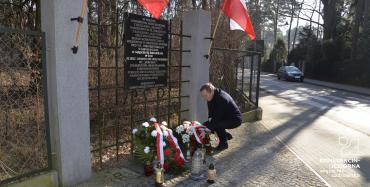 Pośrodku widoczna tablica pamiątkowa umieszczona w ogrodzeniu, po bokach tablicy dwie flagi biało-czerwone, przed tablicą leżą kwiaty i stoją znicze.  Po prawej stronie w przyklęku mężczyzna poprawiający wstążki biało-czerwone w jednej leżących wiązanek.