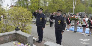 Dwaj przedstawiciele straży miejskiej salutujący przed pomnikiem. W tle uczniowie w biało czerwonych szarfach. 