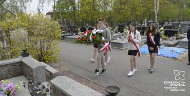 Delegacja uczniów w biało czerwonych szarfach niosąca kwiaty w stronę pomnika. 