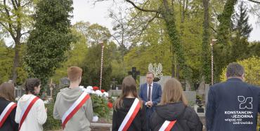 Grupa uczniów z biało czarownymi szarfami stojących z kwiatami przed pomnikiem ofiar drugiej wojny światowej. Przed uczniami stoi przemawiający drugi zastępca burmistrza.  