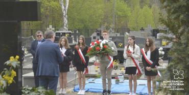 Grupa uczniów w biało czerwonych szarfach z kwiatami. Przed nimi przemawiający Drugi zastępca burmistrza.