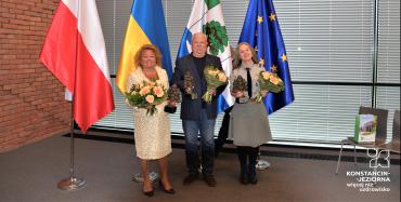 Troje laureatów stoi z kwiatami na tle flag
