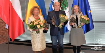 Troje laureatów z kwiatami stoi na tle flag