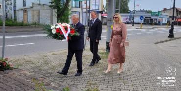 Trzy osoby, przedstawiciele konstancińskiego samorządu, składają kwiaty w miejscu pamięci, osoba w środku trzyma wiązankę kwiatów biało-czerwonych.