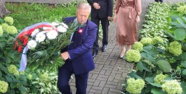 Trzy osoby, przedstawiciele konstancińskiego samorządu, składają kwiaty w miejscu pamięci, osoba w środku trzyma wiązankę kwiatów biało-czerwonych.