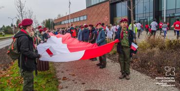 Harcerze pod urzędem trzymają polską flagę, w tle mężczyźni i kobiety ćwiczą przed wystartowaniem w Biegu Niepodległości.