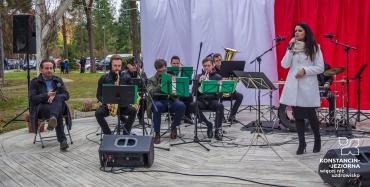 Drewniana scena w parku, na niej siedmiu mężczyzn, którzy grają na instrumentach dętych, obok śpiewa kobieta w białym płaszczyku, z brzegu siedzi mężczyzna z mikrofonem.