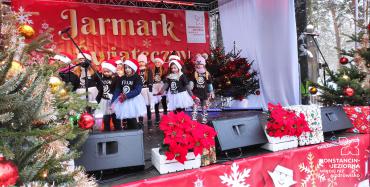 Scena na dworze z napisem Jarmark Świąteczny, na której stoja dzieci. 