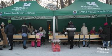 Otwarty namiot z napisem gmina Konstancin-Jeziorn. Pod zadaszeniem przy stole dzieci i kobiety.