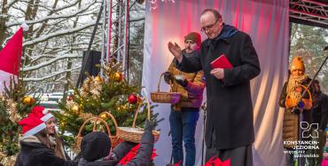 Ksiądz stoi na scenie, ma ręke uciesioną nad koszykami z opłatkami, które trzymają dwie dziewczynki.