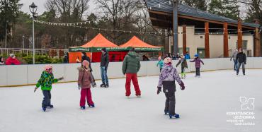 Dzieci i dorośli w czapkach, kaskach i zimowych kurtkach jeździ na łyżwach po sztucznym lodzie. Za nimi drzewa i parkowy amfiteatr.
