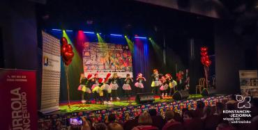 Scena z kolorowymi reflektorami, na niej stoją dziewczynki w czarnych bluzkach, jasnych spódniczkach i mikołajowych czapkach. Przed nimi widownia.