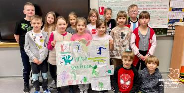 Dziewczyni i chłopcy stoja w sali lekcyjnej, za nimi jest tablica. W rękach trzymają rysunek z napisem: Witamy w Smokolandii.