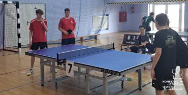 hala sportaowa, na niej stół do tenisa stołowego. A przy nim stoi dwóch chłopców w czerwonych koszulkach i dwóch w czarnych.